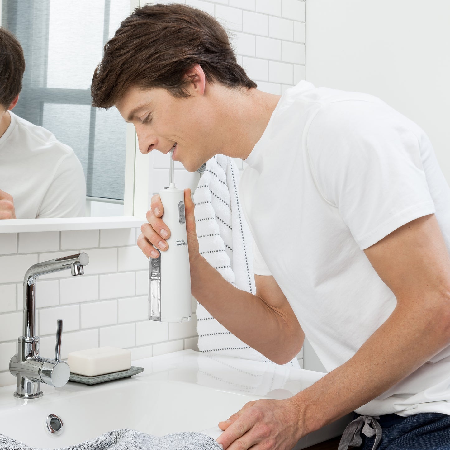 White Cordless Express handheld water flosser in use, with person leaning over the bathroom sink.