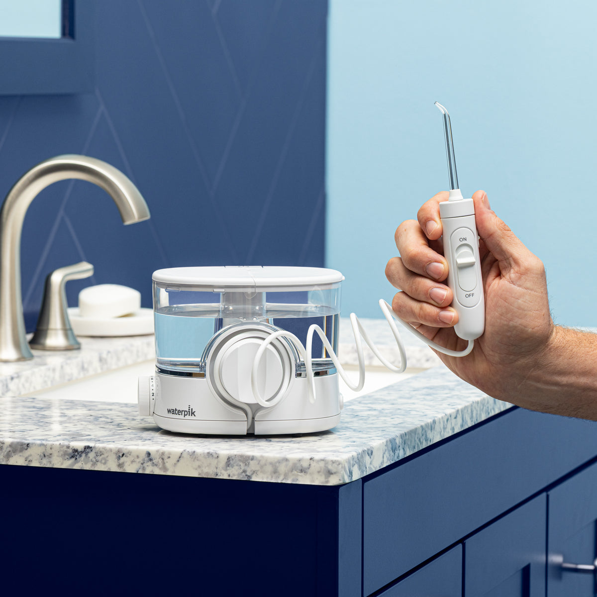 White ION countertop water flosser on a bathroom sink, with hand holding the handle and attached tip, extended by its coiled hose.